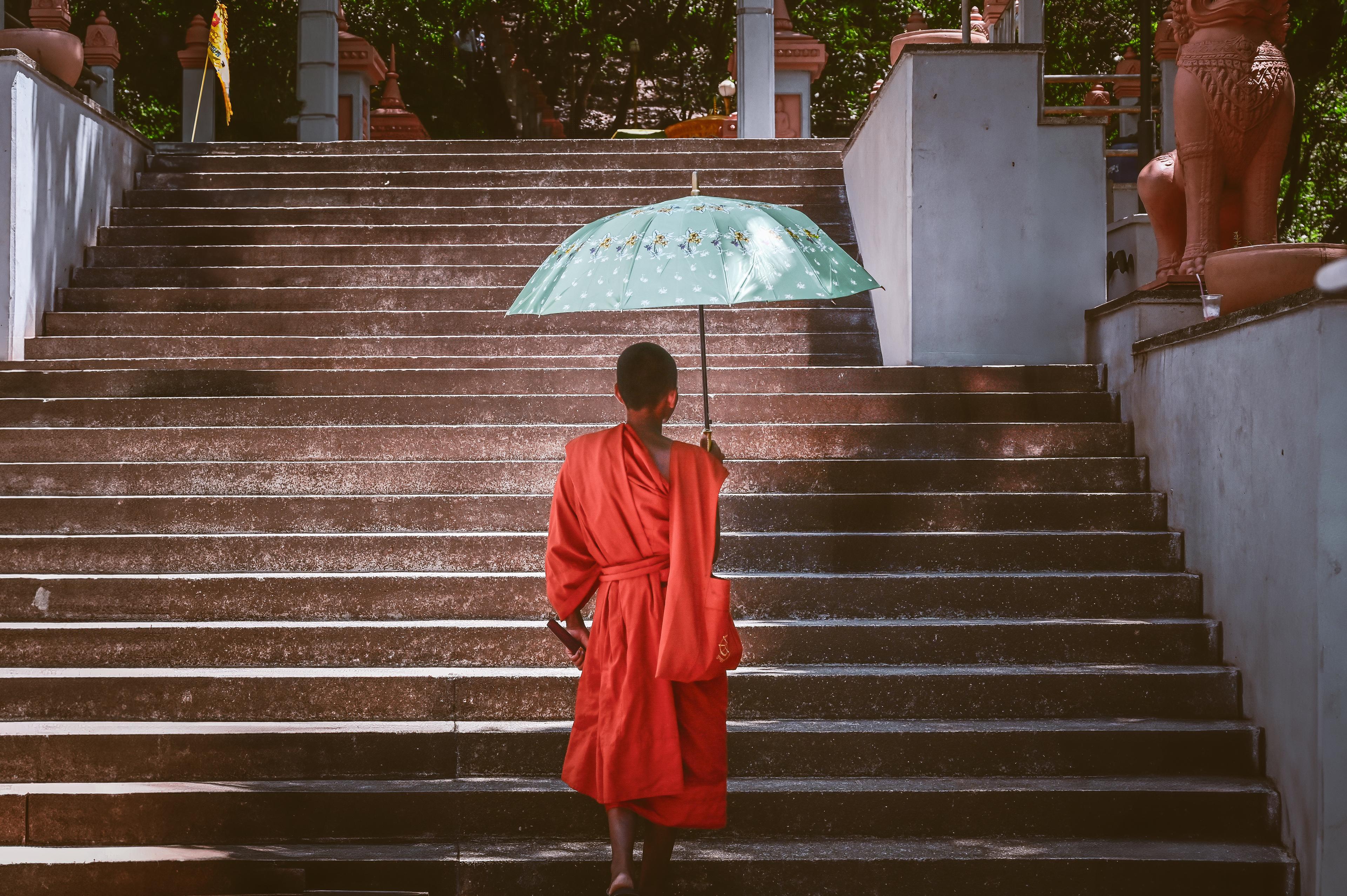 A monk in an orange robe carrying an umbrella up a temple staircase