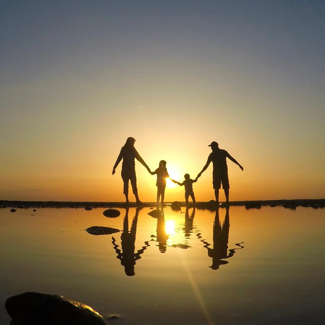 A family of four holding hands, silhouetted against the sunset and reflected in shallow water