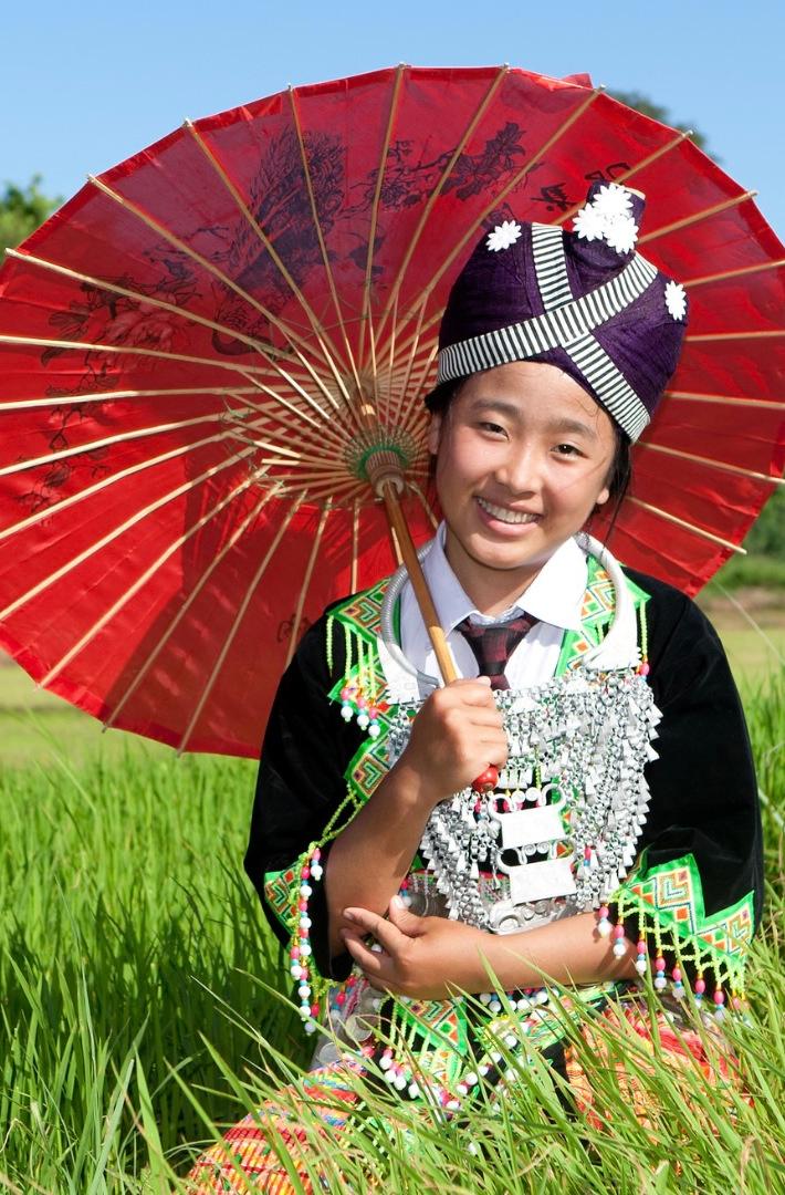 Young woman in Hmong dress holding a red paper parasol in a green rice field