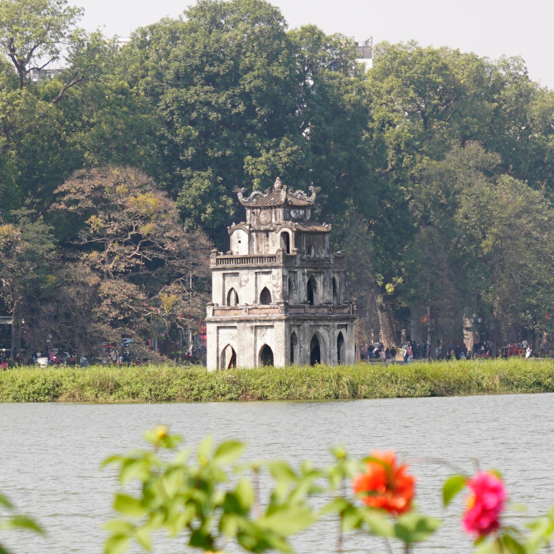 A small stone tower on an island in a city lake, seen through trees and flowers